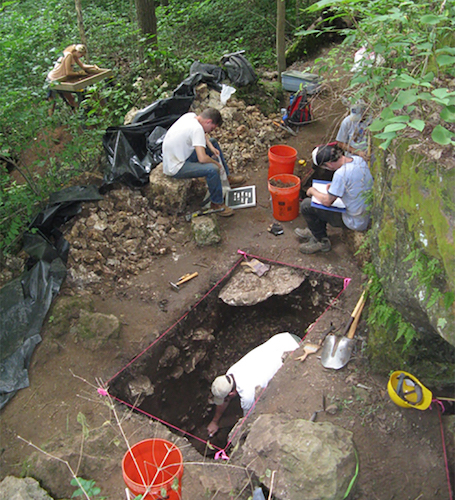 Archaeologist excavate and document a unit in front of a cave entrance.
